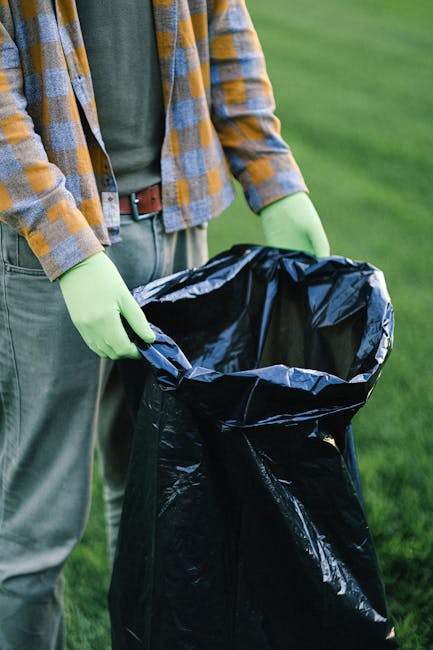 A person wearing orange work overalls and white sneakers stands on a grey paved surface indoors, holding two large blue plastic rubbish bags filled with waste in each hand. The bags appear semi-transl