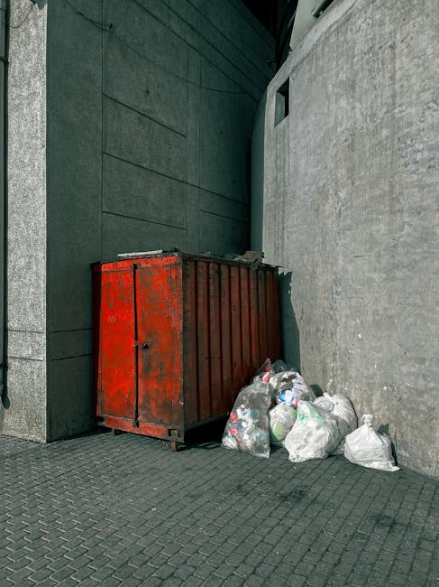 A person wearing a plaid shirt, grey trousers, and light green gloves is holding open a large black plastic rubbish bag outdoors on a grassy area. The bag appears empty, with its top folded over and r